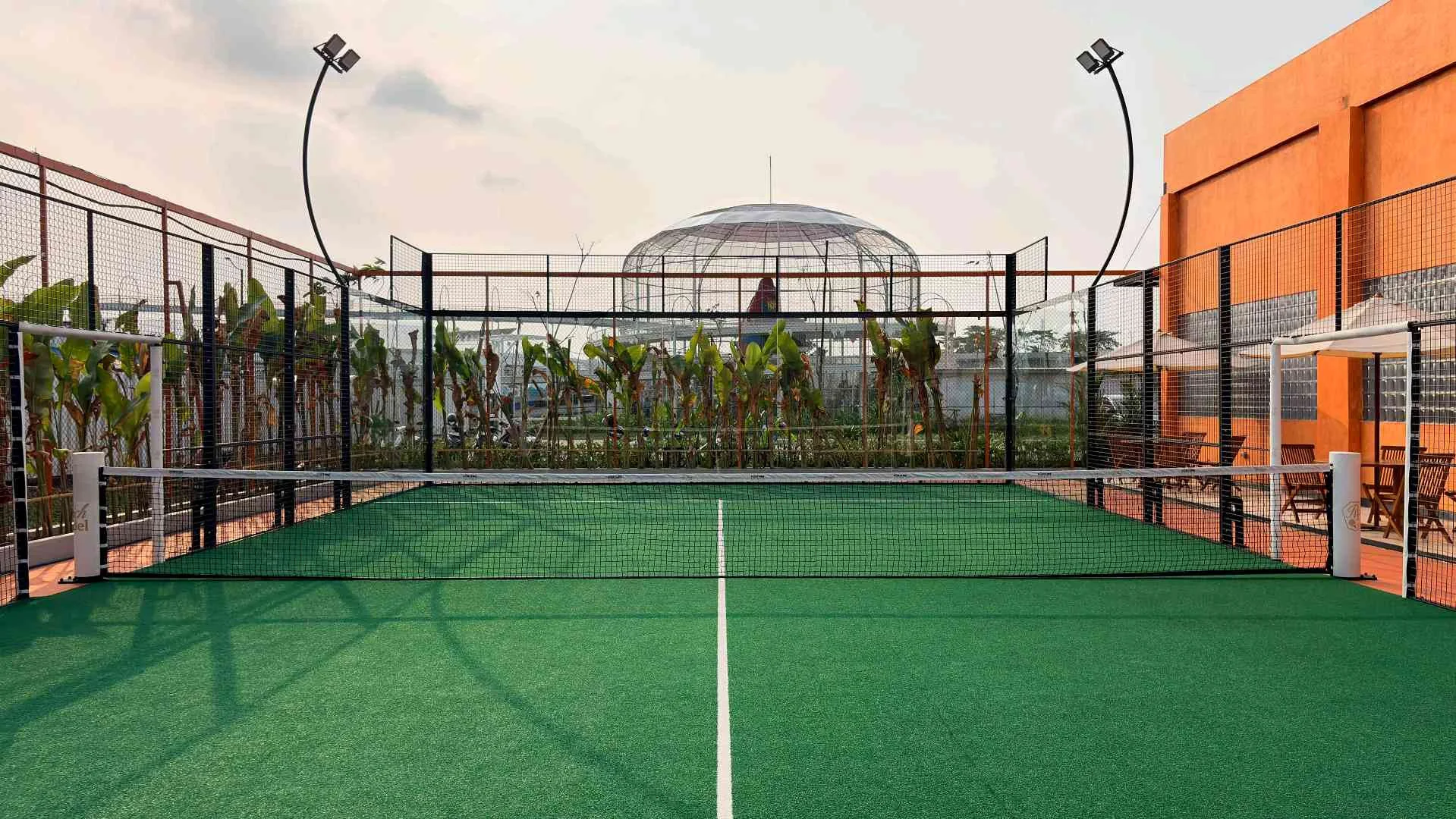 An outdoor court with arched floodlights, banana plants behind the glass and the evening sky
